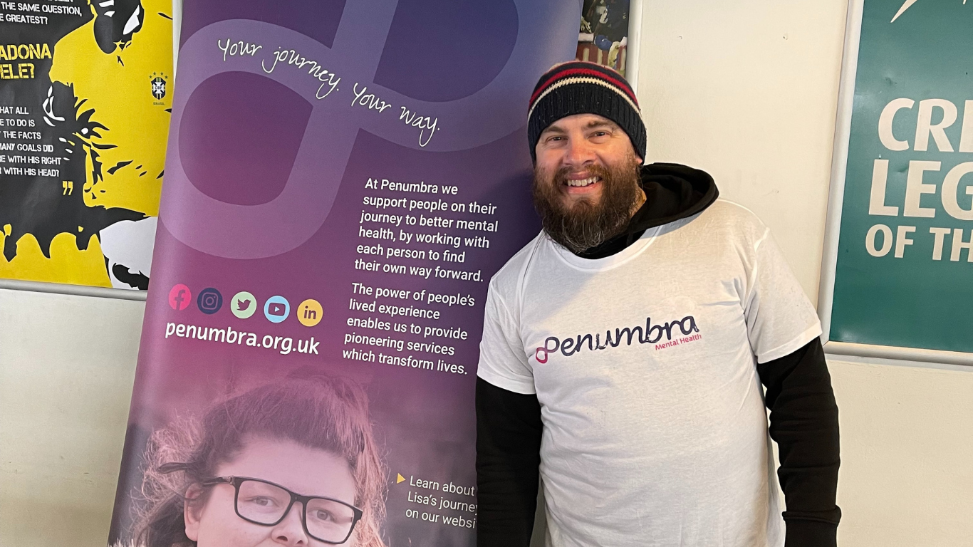 image shows colin smiling to camera wearing his penumbra tee shirt and stanfing beside a penumbra banner
